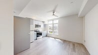 Located in the Derby neighborhood of Seattle, WA, this modern studio offers a thoughtfully designed open-concept layout that maximizes every square foot. Light-toned luxury vinyl plank flooring runs throughout, complementing the clean white walls and creating a bright, cohesive aesthetic.

The kitchen is a standout feature, outfitted with sleek gray cabinetry, open shelving, white countertops, and a full suite of stainless steel appliances including a refrigerator, electric range, over-the-range microwave, and sink. A large picture window positioned adjacent to the kitchen floods the living area with natural light and frames expansive views of the Seattle skyline and surrounding neighborhood rooftops. A ceiling fan overhead provides both comfort and a contemporary design touch.

The living area is open and versatile, offering a clean canvas with recessed lighting and a simple, modern entry door. The bathroom features wood-look flooring consistent with the rest of the unit, a vanity with storage, a toilet, and a walk-in shower with chrome fixtures. The overall design of this studio is clean, functional, and well-suited for urban living in one of Seattle's vibrant neighborhoods.