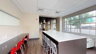 This single-floor commercial space combines open collaborative zones with more intimate seating areas, creating a versatile and well-designed environment. The layout transitions fluidly between distinct functional areas, unified by warm hardwood-style flooring and a neutral color palette of whites, taupes, and dark espresso tones.

A central open area showcases an exposed black ceiling with visible ductwork and structural beams, from which modern pendant lights hang at varying heights, lending an industrial-contemporary character. This zone features lounge seating arranged around a fireplace, with colorful artwork adorning the walls and wire accent chairs adding a modern touch.

A dedicated workspace area includes a substantial dark-base counter with a crisp white surface, lined with sleek metal barstools. Built-in desk space along the perimeter wall provides additional individual work stations, complemented by red accent chairs. Large gridded windows flood this area with natural light and offer views of the surrounding streetscape.

A quieter lounge nook features a pair of white upholstered chairs flanking a round side table, positioned directly beside expansive floor-to-ceiling windows that frame outdoor street views. Throughout, the space balances professional functionality with a welcoming, modern ambiance.