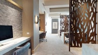 This elegantly designed business center offers a refined and productive environment thoughtfully arranged across a single floor. The space combines open and semi-private work areas, unified by a cohesive palette of warm wood tones, neutral carpeting, and crisp white ceilings with recessed coffers that lend architectural depth overhead.

A defining design element throughout is the use of ornate laser-cut bronze metal screens, which serve as distinctive partitions between workstations. These sculptural dividers provide visual privacy while maintaining an open, airy feel. Individual workstations are outfitted with built-in desks, task chairs, and wall-mounted accent lighting, with textured stone wall treatments adding warmth and character to select areas.

A glass-enclosed conference room serves as a focal point, bathed in abundant natural light from floor-to-ceiling windows that offer views of the surrounding urban landscape. The room features a sleek dark conference table, leather executive chairs, and a contemporary branching chandelier, creating a polished setting for professional meetings.

Smaller semi-private consultation nooks are defined by marble-topped tables set against dramatic dark woven-texture accent walls, ideal for focused one-on-one interactions. The overall ambiance is modern and upscale, blending functional workspaces with carefully curated materials and artistic details that elevate the professional atmosphere throughout.