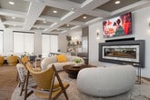 This single-floor club lounge in Meridian, Idaho, offers a generously sized, thoughtfully designed communal space that blends social, recreational, and professional functionality. The entrance greets visitors with soaring double-height ceilings, floor-to-ceiling black-framed glass doors, and herringbone-patterned wood flooring that flows throughout the main areas, creating an immediate sense of openness and sophistication.

The main lounge area serves as the centerpiece, featuring a contemporary linear fireplace, a wall-mounted TV, and a curated seating arrangement with curved cream sofas and mid-century modern accent chairs in warm yellow tones. A coffered ceiling with recessed lighting adds architectural depth overhead. Adjacent to this, a shuffleboard table provides a recreational focal point, while a dining area offers additional seating for gatherings.

A dedicated relaxation room with sage green walls, nature-inspired artwork, and plush lounge seating creates a quieter retreat, also equipped with a wall-mounted TV. A separate conference room with a large table and upholstered seating serves professional needs. The lounge also connects to an exterior fitness facility visible through glass doors. Abundant natural light streams in through large windows throughout, enhancing the inviting, upscale ambiance of this well-appointed community space.