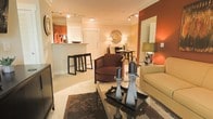 This single-floor residence offers a thoughtfully designed open-concept layout that connects the living, dining, and kitchen areas into one cohesive, flowing space. The living area serves as the heart of the home, featuring warm earth tones — including a rich terracotta accent wall — that create an inviting and cozy ambiance. Plush seating, including a deep brown armchair and a neutral sofa, are arranged around a glass coffee table adorned with decorative candle holders, adding a refined touch to the space. Track lighting runs across the ceiling, providing adjustable illumination that enhances the warm atmosphere throughout. Adjacent to the living space, a compact dining area accommodates an intimate table and chairs, decorated with a sunburst mirror that adds a decorative focal point. The kitchen is accessible via a raised breakfast bar with sleek bar stools, offering both a functional prep space and a casual dining option. Cabinetry and appliances are visible beyond the bar, suggesting a well-equipped culinary space. The open layout, warm color palette, and carefully curated furnishings combine to create a space that feels both comfortable and elegantly appointed, making it well-suited for everyday living and entertaining alike.
