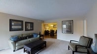 This single-floor residence offers a thoughtfully designed open-concept layout that seamlessly connects the living and dining areas. The living room is a comfortable and inviting space, anchored by a plush sectional sofa with accent pillows, a sleek leather ottoman, and carefully curated wall art in dark frames that add a contemporary touch. Neutral carpet flooring and warm beige walls create a calm, cohesive ambiance throughout. A textured popcorn ceiling adds classic character overhead. The dining area, visible through an open passage, is warmly lit by a chandelier that casts a golden glow over a dark wood dining table and matching chairs. A decorative mirror and accent lamp further enhance the intimate atmosphere of the dining space. Natural light filters into the living area through a window, brightening the space and creating a welcoming environment. The overall design balances comfort and style, with a neutral color palette complemented by rich, dark furnishings. This space is well-suited for both everyday living and entertaining, offering a functional and aesthetically pleasing single-floor layout.