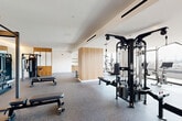 Located at 1070 Bridgeview Way in San Francisco's Mission Bay neighborhood, this modern fitness facility offers a comprehensive and thoughtfully designed workout environment across a single expansive floor. The space is divided into two distinct areas: a well-appointed exercise room and a bright, open studio living area.

The exercise room is generously sized and outfitted with a wide range of professional-grade equipment, including cable machines, a Smith machine, weight benches, dumbbells, kettlebells, rowing machines, and cardio equipment such as treadmills and stationary bikes. Durable rubberized flooring provides safety and functionality throughout. Warm natural wood accent panels and built-in shelving add visual warmth to the otherwise crisp, white interior. Full-length mirrors expand the sense of space, while recessed and linear lighting ensures excellent visibility.

The studio room is a standout feature, showcasing floor-to-ceiling windows and glass doors that flood the light hardwood floors with abundant natural light. A wall of mirrors fitted with elegant wooden ballet barres makes this space ideal for dance, yoga, or barre workouts. The panoramic views of the San Francisco Bay and surrounding cityscape create an inspiring backdrop. A colorful mural adds an artistic touch to the transitional areas, contributing to the facility's dynamic and motivating atmosphere.