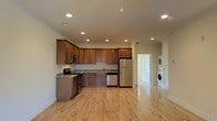 This single-floor residence offers a thoughtfully designed open-concept layout that seamlessly connects the kitchen to the main living area. The kitchen is a standout feature, outfitted with rich warm-toned cabinetry that extends to the ceiling, providing generous storage throughout. Granite countertops add a touch of elegance, complemented by a full suite of stainless steel appliances including a gas range, microwave, dishwasher, and refrigerator. A stainless steel sink is positioned beneath ample counter space, making the kitchen both functional and visually cohesive. Recessed lighting illuminates the space evenly, enhancing the bright and airy atmosphere. Light-toned hardwood flooring runs throughout, adding warmth and continuity to the open floor plan. Crown molding along the ceiling provides a refined architectural detail that elevates the overall aesthetic. A hallway visible from the kitchen leads to additional rooms and notably includes a conveniently located in-unit stacked washer and dryer — a practical amenity that adds significant everyday convenience. The overall ambiance is clean, modern, and move-in ready, making this a highly functional and comfortable living space.