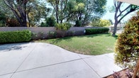 This entrance space offers a welcoming first impression through a combination of well-maintained landscaping and functional outdoor design. A mature tree serves as a natural focal point at the front, providing shade and greenery against the light-colored exterior of the building. Neatly trimmed shrubs, flowering plants, and patches of green lawn line the perimeter, contributing to an established and cared-for appearance.

Smooth concrete walkways curve through the grounds, providing clear and accessible pathways to individual unit entrances. Each unit entrance is distinguished by its own door and short set of steps, offering a sense of privacy and individuality. An exterior staircase with metal railings leads to upper-level access, while a covered carport area accommodates parking conveniently on-site.

The perimeter of the property is defined by a solid block wall, beyond which mature trees rise to create a natural backdrop of lush, verdant canopy. This layering of trees, shrubs, and lawn creates a serene, park-like atmosphere throughout the entrance area. The overall environment is open, airy, and sun-drenched, reflecting a well-organized outdoor setting that balances practical access with pleasant green surroundings.