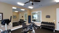This single-floor fitness room provides a practical and well-organized space for exercise. The room features a ceiling fan that promotes air circulation during workouts. Large wall-to-wall mirrors along one side visually expand the space and allow users to monitor their form during exercise. The warm beige walls contribute to an inviting atmosphere, while the dark carpeted flooring adds a grounded, functional feel.

The space is equipped with a selection of strength and cardio equipment, including a stationary bike, a weight bench, and a dumbbell rack stocked with a range of free weights. A large interior window connects the fitness room to an adjacent area, which appears to contain additional cardio equipment such as treadmills, extending the overall workout options available. Bright overhead fluorescent lighting ensures the space is well-lit throughout. A framed piece of artwork on the wall and a mounted plaque add subtle decorative touches. The layout is efficient, allowing users to move between equipment with ease while maintaining a clean, organized environment suitable for a variety of fitness routines.