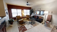 This single-story living space centers around a welcoming living room designed with both comfort and style in mind. A bold, deep red accent wall serves as a striking focal point, contrasting with the neutral tones of the surrounding walls and light-colored carpet underfoot. Multiple large windows allow generous natural light to fill the room, creating a bright and open atmosphere.

The seating arrangement includes a dark leather sofa adorned with richly colored throw pillows in earthy tones, complemented by a pair of warm brown upholstered armchairs. A matching ottoman adds additional comfort and flexibility to the layout. Anchoring the seating area is a large patchwork-style area rug featuring a blend of red, blue, gray, and cream tones, adding visual depth and texture to the space.

A glass-top coffee table with ornate metal scrollwork sits at the center of the arrangement, echoing the decorative iron side table nearby. A ceiling fan overhead provides both functionality and subtle style. A rustic wood-framed mirror mounted on one wall adds dimension, while a tall indoor plant introduces a natural, organic element. The overall ambiance is warm, layered, and thoughtfully curated.