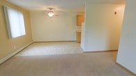 This single-floor residence offers a functional and straightforward layout with a clean, neutral aesthetic throughout. The living area features plush, light-colored carpeting that adds warmth underfoot, while the adjacent dining and kitchen zone transitions seamlessly to light-toned tile flooring. A ceiling fan with an integrated light fixture is centrally mounted overhead, providing both ventilation and ambient lighting to the open living space. The kitchen is positioned in an open-concept fashion, partially separated by a partial wall that creates a natural division without sacrificing the sense of openness. Natural wood-toned cabinetry lines the upper kitchen area, complementing the light walls and overall neutral color palette. A full-size refrigerator anchors the kitchen, which benefits from the same bright, airy atmosphere found throughout the space. The walls are finished in a soft, off-white tone that reflects light and contributes to the spacious feel of the interior. Baseboard heating runs along the perimeter of the tile area, ensuring efficient warmth distribution. The overall ambiance is calm and understated, offering a blank canvas that lends itself well to a variety of interior styles and furnishing preferences.