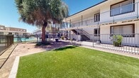 Located at 4235 N 35th Ave in Phoenix, Arizona, this outdoor common area serves as a dedicated pet-friendly amenity space within a multi-unit residential community. The space is fully enclosed by a sturdy iron perimeter fence, providing a secure environment for pets and their owners. A central feature of the area is a well-maintained patch of lush artificial turf, offering a clean and comfortable surface for recreation. Mature trees, including a prominent large-canopied tree near the center, provide natural shade — a particularly valuable asset under the Arizona sun. The surrounding ground is finished with natural compacted gravel, and designated waste stations are conveniently positioned throughout the enclosure. The two-story residential building frames the space on multiple sides, with covered walkways, private balconies, and exterior staircases visible along the facade. The building's white and neutral-toned exterior with dark metal railings lends a clean, well-maintained appearance. Under the clear blue Phoenix sky, the space feels open and airy, offering residents and their pets a comfortable, functional outdoor retreat that balances utility with a welcoming atmosphere.