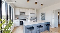 Located in the heart of Durham, NC, this thoughtfully designed 2-bedroom, 2-bathroom residence offers a modern aesthetic with clean lines and quality finishes throughout. The open-concept kitchen and living area serves as the centerpiece of the home, where a sleek kitchen features white cabinetry, stainless steel appliances, a subway tile backsplash, and a striking blue island with a quartz countertop and bar seating. Light wood flooring flows seamlessly throughout the space, enhancing the bright and airy atmosphere. The living room is bathed in natural light from expansive black-framed windows that frame views of the vibrant Durham cityscape, with direct access to a private balcony. The primary bedroom continues this connection to the outdoors via sliding glass doors to the balcony, offering urban views and light wood flooring. It includes a well-appointed closet with wire shelving. The second bedroom is equally bright, featuring its own closet and a crisp, neutral palette. Both bathrooms are finished with marble-topped floating vanities, white subway tile tub-shower combinations, and modern globe light fixtures set against cool gray walls. Three closets total provide ample storage throughout this efficiently designed and stylish urban residence.