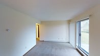 This single-floor space presents a clean, minimalist living area characterized by its neutral color palette and straightforward layout. The room features wall-to-wall carpeting in a light gray tone, contributing to a quiet and comfortable atmosphere underfoot. Smooth, white ceilings and cream-colored walls create a bright, airy feel throughout the space. A baseboard heating unit runs along the far wall, providing an efficient and unobtrusive heating solution. Natural light enters through a large window fitted with vertical blinds on the right side of the room, allowing for adjustable light control. A doorway visible in the background connects to an adjacent area, suggesting a functional flow between rooms. The overall design is simple and practical, with ample open floor space that offers flexibility for a variety of furniture arrangements. The clean walls, neutral finishes, and understated details make this a versatile space well-suited for customization to suit personal style preferences.
