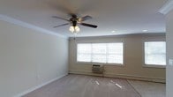 This single-floor residential space offers a clean and functional layout designed for comfortable everyday living. The main living area is finished with wall-to-wall carpeting that adds warmth underfoot, while crisp white walls and crown molding lend a classic, polished character to the interior. A ceiling fan with an integrated light fixture is centrally mounted, providing both ambient lighting and year-round air circulation throughout the room. A wide window dressed with horizontal blinds allows natural light to filter in, brightening the space while offering privacy control. Beneath the window, a through-the-wall air conditioning and heating unit ensures climate comfort in all seasons, complemented by baseboard heating elements that run along the perimeter walls. Recessed lighting in the ceiling adds an additional layer of illumination, contributing to a well-lit and inviting atmosphere. The neutral color palette and understated finishes create a versatile backdrop, making the space easy to personalize and furnish to suit a variety of preferences. Overall, the space presents a practical and move-in ready environment with thoughtful climate control features and timeless architectural detailing.