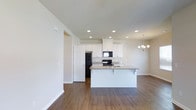 This single-story home showcases a well-designed open-concept layout that seamlessly connects the kitchen and living areas, creating a spacious and functional environment. The kitchen is a standout feature, outfitted with crisp white shaker-style cabinetry that extends to the upper walls, providing generous storage. A granite-topped island with a breakfast bar overhang serves as both a prep space and casual dining area, anchoring the kitchen within the open floor plan. Black appliances, including a refrigerator, range, and over-the-range microwave, offer a clean contrast against the white cabinetry. Recessed lighting is installed throughout the ceiling, ensuring consistent, well-distributed illumination across the space. An elegant chandelier is visible in the adjacent dining or living area, adding a refined decorative touch. Rich, dark hardwood-style flooring runs continuously throughout, grounding the space with warmth and visual cohesion. The neutral white walls and clean lines contribute to a bright, modern aesthetic. A interior door is accessible from the kitchen area, suggesting convenient connectivity to additional rooms within the home.