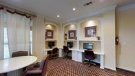 This single-floor business center is thoughtfully designed to provide a functional and comfortable workspace. The room's most distinctive architectural feature is a trio of arched built-in alcoves, each housing a dedicated workstation complete with a desktop computer, a built-in white desk surface, and overhead accent lighting. The alcoves are framed in crisp white millwork and accented with decorative framed artwork, lending a polished, professional atmosphere to the space.

Crown molding runs along the ceiling line, adding a refined architectural detail, while recessed lighting provides consistent, even illumination throughout. The rich, plaid-patterned carpet in deep burgundy and tan tones grounds the room with warmth and texture. Each workstation is paired with a high-back office chair upholstered in dark burgundy fabric, ensuring comfort during extended use.

Natural light filters in through windows fitted with blinds, offering a pleasant connection to the outdoors while maintaining privacy and glare control. A seating area with upholstered chairs and a round table is visible at the edge of the room, providing an additional area for casual meetings or reading. Overall, the space balances practicality with classic elegance, making it a well-equipped and inviting environment for productivity.