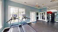 Located at Enterprise Way in Maryville, Tennessee, this fitness center offers a well-appointed, single-story space designed to support a comprehensive workout experience. The interior is finished with soft blue walls accented by crisp white crown molding, creating a clean and inviting atmosphere throughout. Durable rubber flooring covers the workout areas, providing both safety and functionality for a range of exercises.

The space is divided into distinct workout zones. One area is dedicated to cardio equipment, housing multiple treadmills and elliptical trainers from TRUE Fitness, positioned to take advantage of the abundant natural light streaming through large, white-framed windows that overlook the surrounding landscaping. Ceiling fans overhead contribute to comfort during workouts. A second area accommodates strength training, featuring a cable machine, adjustable weight bench, and a dumbbell rack, with a full-length wall mirror enhancing the sense of space and allowing users to monitor their form.

Additional amenities include a water station near the entrance and glass-panel doors that connect sections of the facility. Recessed lighting supplements the natural light, ensuring the space remains bright throughout the day. The overall ambiance is functional yet comfortable, offering users a well-rounded environment for fitness.