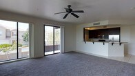 This single-floor space offers an open, airy layout designed for comfortable everyday living. The main living area is finished with plush wall-to-wall carpeting and neutral-toned walls that create a clean, welcoming ambiance. A ceiling fan centered overhead provides both style and functionality, ensuring year-round comfort. A large sliding glass door floods the room with natural light and opens directly onto a private balcony, offering pleasant outdoor views of surrounding greenery — a standout feature of the space.

The kitchen is thoughtfully integrated into the layout via a pass-through design, featuring dark espresso-toned cabinetry, a light granite-style countertop with a breakfast bar overhang ideal for casual dining, and stainless steel appliances including a microwave. Overhead recessed lighting illuminates the kitchen workspace efficiently. A utility sink and ample cabinetry provide practical storage and functionality.

The overall design emphasizes an open flow between the kitchen and living areas, making the space feel expansive and well-connected. With its combination of natural light, outdoor access, and modern kitchen finishes, this space strikes a balance between comfort and practicality.