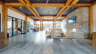The 53 West Exchange Clubhouse in Gainesville, Georgia, is a single-story community facility that masterfully combines rustic warmth with contemporary industrial design. The structure is defined by prominent exposed wood beam ceilings, accented by dark corrugated metal panels and recessed lighting, creating a striking architectural contrast throughout the interior.

The main gathering area is expansive and open, featuring polished concrete floors that reflect natural light flooding in from large glass doors and open-air passages. A statement chandelier anchors the central lounge, where wood-framed seating is arranged around a stone accent wall housing a fireplace, contributing to a welcoming, resort-like ambiance.

The clubhouse flows seamlessly to a covered outdoor patio, framed by heavy timber posts and strung with Edison bulb lighting, providing views toward the pool area and surrounding community. A semi-enclosed entry vestibule with built-in wood benches offers a casual seating nook with open views to the outdoors.

Inside, a bar counter with a live-edge wood top and wood stools serves as a social hub, complemented by wall-mounted TVs. Additional areas feature patterned carpet, shiplap walls, and bistro-style seating, offering versatile spaces for both relaxed gatherings and community events.