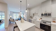 This single-floor residence offers a thoughtfully designed open-concept layout that seamlessly connects the kitchen, dining, and living areas. The kitchen is a standout feature, outfitted with light gray shaker-style cabinetry, white quartz countertops, a full suite of stainless steel appliances — including a refrigerator, range, microwave, and dishwasher — and a generous central island illuminated by stylish pendant lighting. Light wood-tone flooring flows consistently throughout the space, contributing to a clean, cohesive aesthetic.

The living area is bright and welcoming, benefiting from a large window that invites natural light and frames views of the surrounding greenery. The space comfortably accommodates a sectional sofa and coffee table, with pendant and floor lighting adding warmth to the neutral palette.

The bedroom is a serene retreat, featuring large windows with outdoor views, light-filtering roller shades, and rich drape panels. It offers ample space for a full bedroom suite and provides a calm, restful atmosphere.

The bathroom is finished with light wood-look flooring, a glass-enclosed walk-in shower with gray tile surround, a vanity with integrated sink, and open wall shelving — combining functionality with a clean, contemporary design.