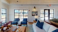 This single-story clubhouse showcases a refined coastal farmhouse design that seamlessly blends rustic warmth with modern elegance. The interior is defined by striking white shiplap walls, rich hardwood floors laid in a herringbone pattern in select areas, and soaring vaulted ceilings finished in warm natural wood — creating an inviting, airy atmosphere throughout.

The main gathering area serves as the heart of the space, anchored by a whitewashed brick fireplace with a reclaimed wood mantel and a mounted television above. Comfortable seating in navy blue and neutral tones is arranged around a wooden coffee table, making it an ideal spot for relaxed socializing. Large black-framed windows and glass doors flood the room with natural light and offer views of the surrounding landscape.

An expansive dining and event area features multiple dining tables with navy-accented bases and woven chairs, illuminated by statement wagon-wheel chandeliers and industrial-style wall sconces. A dedicated service bar area adds functionality, showcasing deep green cabinetry, a stone countertop, a sink, and open reclaimed wood shelving — a tasteful focal point that bridges style and practicality. Coastal-themed artwork and eclectic décor details complete the character of this well-appointed communal space.