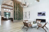 This single-story community clubhouse in Owens Cross Roads, Alabama, showcases a thoughtfully designed interior that blends natural materials with modern aesthetics. The expansive open floor plan is anchored by soaring vaulted ceilings accented with exposed wood beams, creating an airy, welcoming atmosphere throughout.

A standout architectural feature is the striking vertical wood-slat divider wrapped in lush trailing greenery, serving as a natural partition between zones while adding a botanical element to the interior. Light hardwood flooring flows continuously throughout the space, complementing the white walls and warm wood tones.

The main gathering area features multiple seating arrangements, including a large communal dining table beneath a statement chandelier, a lounge with a live-edge wood bar counter, and a relaxed TV lounge with floor-to-ceiling windows that draw in abundant natural light and outdoor views. An arched alcove housing a mounted display screen adds an elegant architectural detail.

A separate lounge area with exposed beam ceilings connects to an outdoor patio through sliding glass doors. A dedicated business center offers workstations beneath industrial pendant lighting with garden-facing windows. A cozy banquette nook with tufted velvet seating and a game table rounds out the amenities, offering a casual retreat within the clubhouse.