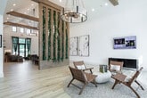 This single-story community clubhouse in Owens Cross Roads, Alabama, showcases a thoughtfully designed interior that blends natural materials with modern aesthetics. The expansive open floor plan is anchored by soaring vaulted ceilings accented with exposed wood beams, creating an airy, welcoming atmosphere throughout.

A standout architectural feature is the striking vertical wood-slat divider wrapped in lush trailing greenery, serving as a natural partition between zones while adding a botanical element to the interior. Light hardwood flooring flows continuously throughout the space, complementing the white walls and warm wood tones.

The main gathering area features multiple seating arrangements, including a large communal dining table beneath a statement chandelier, a lounge with a live-edge wood bar counter, and a relaxed TV lounge with floor-to-ceiling windows that draw in abundant natural light and outdoor views. An arched alcove housing a mounted display screen adds an elegant architectural detail.

A separate lounge area with exposed beam ceilings connects to an outdoor patio through sliding glass doors. A dedicated business center offers workstations beneath industrial pendant lighting with garden-facing windows. A cozy banquette nook with tufted velvet seating and a game table rounds out the amenities, offering a casual retreat within the clubhouse.