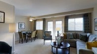 This single-floor residence offers a well-designed open-plan layout that seamlessly connects the living and dining areas into one cohesive, functional space. The living room is anchored by a plush sofa adorned with teal and mustard accent pillows, complemented by a classic round dark-wood coffee table and an elegant accent chair with a patterned cushion. Natural light floods the interior through multiple large windows fitted with white blinds and coordinating blue-and-gold patterned drapes, creating a warm and welcoming atmosphere. A ceiling fan with a light fixture adds both comfort and a refined touch to the room. The adjacent dining area accommodates a full dining table surrounded by upholstered chairs in muted olive and taupe tones, making it ideal for everyday meals or entertaining guests. Neutral-toned carpeting runs throughout, contributing to the space's cohesive and calming aesthetic. The walls are finished in a warm beige, enhancing the overall sense of warmth. French doors open to the exterior, inviting in additional natural light and providing a seamless connection to the outdoors. The space strikes a balance between comfort and style, making it an inviting and practical home environment.
