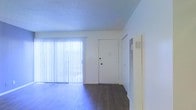 This two-floor space offers a clean, functional layout designed to maximize comfort and usability. The main living area immediately captures attention with its rich, dark hardwood-style flooring, which contrasts neatly against crisp white walls. A large sliding glass door, dressed with vertical blinds, floods the room with natural light and provides direct access to an outdoor area, enhancing the sense of openness throughout the space. A textured ceiling adds a classic architectural touch overhead. A built-in HVAC vent is integrated into the wall, ensuring climate control is both practical and unobtrusive. The front entry door is centrally positioned, offering a straightforward and welcoming point of arrival. The overall aesthetic is clean and neutral, providing a versatile canvas well-suited to a variety of interior styles. Spanning two floors, the space provides ample room for distinct living, sleeping, and utility areas across its levels. The open feel of the main living area, combined with the abundance of natural light and simple, well-maintained finishes, contributes to a bright and inviting ambiance throughout the property.