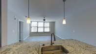 This single-floor space showcases an open-concept layout that seamlessly connects the kitchen and living area. The kitchen is anchored by a dark-cabinetry island topped with light granite countertops, complete with an undermount stainless steel sink and a sleek gooseneck faucet. Two pendant lights with frosted glass shades hang above the island, providing focused task lighting that complements the flush-mount ceiling fixture nearby. The cabinetry's rich, dark wood finish creates a striking contrast against the light granite and the neutral white walls throughout. Hardwood-style flooring extends continuously from the kitchen into the open living area, enhancing the sense of flow and cohesion. The living space is bright and airy, with a large window dressed in horizontal blinds that allows generous natural light to fill the room. A ceiling fan centered in the living area offers both functionality and a finishing touch to the space. Clean white baseboards and smooth walls contribute to a crisp, modern aesthetic throughout, while a hallway visible in the background suggests additional rooms beyond the main open-concept area.