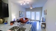 This single-floor space is designed with an open-concept layout that seamlessly connects the kitchen and living area, creating a cohesive and functional environment. The kitchen is appointed with sleek granite countertops and a double-basin sink, offering both style and practicality. Track lighting overhead adds focused illumination, complementing the clean, neutral tones of the space.

The living area is warm and inviting, anchored by a rich terracotta-toned sofa and accented with a bold blue-striped area rug that adds a pop of color to the light-toned flooring. A ceiling fan with an integrated light fixture ensures year-round comfort. A striking large-format wave artwork serves as a focal point on one wall, lending an artistic character to the room.

French doors along the far wall flood the space with natural light while providing access to the outdoors, enhancing the airy, open feel of the interior. Bright white walls and light-colored floors further amplify the sense of openness throughout. The living room also features a flat-screen TV and entertainment setup, making the space as functional as it is aesthetically pleasing. Overall, the unit offers a bright, comfortable, and well-appointed living environment.