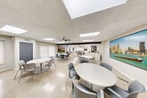Located at 25225 Greenfield Rd in Southfield, Michigan, this single-floor multipurpose facility offers a well-organized and versatile layout designed for communal use. The main open area is generously proportioned, featuring polished terrazzo flooring throughout that lends a clean, durable aesthetic to the entire space. Recessed panel ceilings with integrated skylight panels allow abundant natural light to filter in, complementing the ceiling fans that promote comfortable air circulation.

The gathering area is divided into distinct functional zones. A dining section features multiple round white tables paired with modern gray chairs, accommodating a variety of group sizes. A separate lounge zone is furnished with plush gray modular seating arranged around a central coffee table, along with a wall-mounted television display — ideal for relaxed gatherings or presentations. A standalone upright piano adds a cultural and recreational touch to the space.

Large framed photographic artwork adorning the walls reflects a community-oriented character, with images depicting local and historical landmarks. Roller shades on the windows provide adjustable light control throughout.

The kitchen is efficiently designed with white shaker cabinetry, marble-look quartz countertops, a textured tile backsplash, stainless steel appliances including a refrigerator, and wood-look flooring — offering full functionality for catering or event preparation. Multiple gray access doors connect the various zones, ensuring smooth circulation throughout this well-appointed community space.