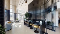 This contemporary residential lobby makes an immediate impression with its soaring double-height ceilings and expansive floor-to-ceiling glass curtain wall that floods the interior with abundant natural light and offers sweeping views of the surrounding streetscape. Structural columns clad in deep navy tile add a striking architectural contrast against the light-toned porcelain tile flooring, which stretches seamlessly throughout the space.

The main lounge area is thoughtfully appointed with an eclectic mix of modern seating — rounded swivel chairs, a plush sofa, and marble-topped accent tables — arranged atop patterned area rugs to create intimate conversation zones. Lush potted greenery and large indoor plants are strategically placed throughout, bringing a natural, refreshing energy to the interior.

A sleek co-working bar lined with upholstered stools runs along a dramatic floor-to-ceiling slatted black partition, offering a dedicated workspace within the lobby. Adjacent to this is a well-equipped beverage station framed by warm light wood cabinetry and a geometric patterned tile backsplash. The mailroom area features a built-in bank of mailboxes set into a wood-paneled surround. Elevator access and additional corridors connect seamlessly, completing this polished and functional ground-floor amenity space.