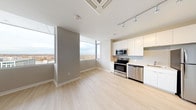 This single-floor unit presents a clean, contemporary design throughout its open-concept layout. The living and dining area is characterized by light hardwood flooring, neutral gray walls, and large floor-to-ceiling windows that frame sweeping outdoor views of the surrounding treetops and skyline, flooding the space with natural light. A ceiling-mounted HVAC unit and track lighting enhance both comfort and functionality in this area.

The kitchen is efficiently designed with white flat-front cabinetry extending to the ceiling, light countertops, and a full suite of stainless steel appliances, including a refrigerator, range, built-in microwave, and dishwasher. A subway tile backsplash adds a subtle design detail to the culinary space.

The bedroom features the same light hardwood flooring, a ceiling-mounted HVAC unit, and a walk-in closet outfitted with wire shelving for practical storage. A secondary carpeted room with neutral gray walls provides additional flexible living space, complete with a utility panel.

The bathroom is finished in a clean, all-white palette, featuring large-format floor tiles, a soaking tub with shower, white vanity cabinetry, and chrome fixtures, contributing to the unit's cohesive, modern aesthetic.