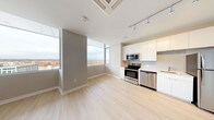 This single-floor unit presents a clean, contemporary design throughout its open-concept layout. The living and dining area is characterized by light hardwood flooring, neutral gray walls, and large floor-to-ceiling windows that frame sweeping outdoor views of the surrounding treetops and skyline, flooding the space with natural light. A ceiling-mounted HVAC unit and track lighting enhance both comfort and functionality in this area.

The kitchen is efficiently designed with white flat-front cabinetry extending to the ceiling, light countertops, and a full suite of stainless steel appliances, including a refrigerator, range, built-in microwave, and dishwasher. A subway tile backsplash adds a subtle design detail to the culinary space.

The bedroom features the same light hardwood flooring, a ceiling-mounted HVAC unit, and a walk-in closet outfitted with wire shelving for practical storage. A secondary carpeted room with neutral gray walls provides additional flexible living space, complete with a utility panel.

The bathroom is finished in a clean, all-white palette, featuring large-format floor tiles, a soaking tub with shower, white vanity cabinetry, and chrome fixtures, contributing to the unit's cohesive, modern aesthetic.