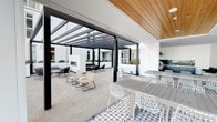 Located at 13448 Beach Ave in the Mar Vista neighborhood of Los Angeles, this outdoor amenity level offers a thoughtfully designed retreat that blends modern architecture with Southern California's indoor-outdoor lifestyle. The space is anchored by a resort-style swimming pool surrounded by a generous sun terrace, furnished with sleek chaise lounges and teak-framed seating with deep cushions, ideal for relaxing under the clear Los Angeles sky. Patio heaters are positioned throughout the deck, extending the usability of the space year-round. A covered outdoor lounge area provides a sophisticated transition between open-air and sheltered environments, featuring a striking pergola with dark steel framing and warm wood-clad ceilings fitted with recessed lighting. Beneath the pergola, the space is divided into distinct zones: communal dining tables with white wire chairs sit alongside a casual lounge arrangement centered around a freestanding fireplace. Geometric patterned tile flooring adds visual interest and delineates different seating areas. The surrounding white modern building rises with clean lines and private balconies, while views of the Los Angeles skyline and iconic palm trees frame the horizon, reinforcing the quintessential California ambiance of this well-appointed outdoor space.