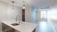 Located at 90 State St in the heart of Albany, NY, this efficiently designed single-floor unit offers a thoughtfully arranged layout comprising a kitchen, living area, bathroom, hallway, and closet. The kitchen is a clear standout, showcasing an expansive waterfall-edge quartz island with an undermount stainless steel sink and a high-arc pull-down faucet. Rich, dark-toned cabinetry lines the walls, complemented by a classic white subway tile backsplash, stainless steel appliances including a professional-grade gas range, built-in microwave, and a full-size refrigerator. Pendant lighting with exposed Edison bulbs and track lighting enhance the kitchen's contemporary aesthetic. The living area features light wood-tone flooring, neutral walls, and a window that invites natural light into the space. The bathroom is finished with marble-look tile flooring, a dark-cabinetry vanity with an integrated sink, a wall-mounted mirror, and a glass-enclosed walk-in shower clad in white subway tile with built-in shelving niches. Throughout the unit, clean lines, neutral tones, and modern fixtures combine to create a cohesive, urban-contemporary ambiance well-suited to downtown Albany living.