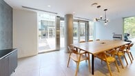 Located at 1770 Pendrell Street in Vancouver's West End, this single-floor dining amenity room offers a thoughtfully designed gathering space that balances modern aesthetics with practical functionality. Large floor-to-ceiling glass doors and windows flood the interior with abundant natural light while providing open views of the surrounding outdoor courtyard and lush greenery, creating a seamless connection between inside and out.

The space is anchored by a generously sized rectangular dining table crafted in warm natural wood, surrounded by a set of contemporary Scandinavian-style chairs in a complementary honey tone. Overhead, a sculptural multi-globe pendant light fixture adds an artful focal point and warm ambient illumination. Large-format light grey porcelain tiles span the floor throughout, contributing to the room's clean, open feel.

A full-length built-in kitchenette lines one wall, featuring warm oak cabinetry, a stainless steel built-in oven, a sink with a matte black faucet, and upper white cabinetry with under-cabinet lighting. A striking accent wall finished in a textured teal patterned wallcovering serves as a media wall, complete with a wall-mounted flat-screen television and a sleek dark sideboard beneath. Exposed concrete columns add an industrial architectural character that complements the refined modern interior design throughout.