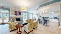 Located in Morehead City, NC, this expansive single-floor community clubhouse offers a thoughtfully designed amenity space with a coastal-inspired aesthetic. The main lounge is a welcoming gathering area anchored by a striking stone fireplace, complemented by light hardwood-style flooring, crown molding, and soft teal accent walls. Large windows and French doors flood the space with natural light while providing views of the surrounding outdoor areas. A comfortable seating area with patterned armchairs invites relaxation, while an adjacent dining nook with a round table and modern chairs offers a more intimate setting. The full kitchen is well-appointed with white shaker cabinetry, granite countertops, a vibrant blue glass tile backsplash, stainless steel appliances, and a peninsula with bar seating — ideal for community gatherings and events. A bright, sunlit lounge room features tufted sofas, elegant drapery, and refined décor with a nautical nod. The fitness center is generously sized and divided into two areas: one dedicated to free weights, a weight bench, and exercise mats, and another equipped with spin bikes and a functional training rig with a punching bag. Two bathrooms and a hallway round out this well-appointed community facility.