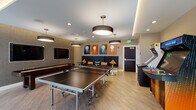 This single-floor recreational game room is thoughtfully designed to offer a dynamic and engaging entertainment experience. The space features light wood-style flooring throughout, creating a clean, modern foundation that ties the room together. Recessed lighting is complemented by a series of bold pendant fixtures with dark drum shades, providing warm, even illumination across the entire area.

At the center of the room, a full-size ping-pong table serves as the focal point, while a foosball table and a billiards table offer additional gameplay options. A dedicated arcade corner houses two classic upright arcade cabinets, adding a nostalgic touch to the contemporary setting.

A built-in lounge bench upholstered in deep navy blue runs along one wall, adorned with mustard yellow and patterned accent pillows. Above it, a series of four bold, modern art panels featuring abstract circular motifs in shades of blue, orange, and gold add a striking visual element. Circular high-top tables with sleek pedestal stools are positioned throughout for casual seating.

A wall-mounted flat-screen television is set against a textured accent wall, creating a dedicated media area. Large glass doors on one side of the room invite natural light and provide access to an adjacent space. The overall ambiance is modern, lively, and well-appointed for social recreation.