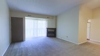 This single-floor space offers a clean, functional layout designed for comfortable everyday living. The main living area is finished with wall-to-wall carpeting in a neutral tone, creating a soft and inviting atmosphere throughout. A standout feature of the room is the corner fireplace, framed in dark wood paneling, which serves as a focal point and adds a cozy, practical element to the space. Adjacent to the fireplace, a large sliding glass door fitted with vertical blinds allows an abundance of natural light to pour into the room, brightening the interior and offering the option for easy indoor-outdoor access. The walls are finished in a light, neutral palette that enhances the sense of openness, while the ceiling is generously proportioned, contributing to an airy feel. Overhead HVAC vents are integrated into both the ceiling and upper wall, ensuring climate control throughout the space. The overall ambiance is calm and understated, providing a versatile blank canvas well-suited for a variety of furnishing and design styles.