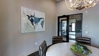 This single-floor space exudes a refined, contemporary aesthetic with a warm Western-inspired character. The interior is anchored by a well-appointed meeting or lounge area, where a round marble-top table is surrounded by rich brown leather chairs, creating an inviting atmosphere for gatherings or collaboration. A striking large-format watercolor painting of a longhorn steer serves as a bold focal point on the soft gray walls, adding a distinctive artistic touch that defines the space's personality.

Overhead, an antler chandelier introduces a rustic yet elegant design element that complements the space's upscale ambiance. The neutral gray wall palette provides a sophisticated backdrop that allows the curated furnishings and artwork to take center stage.

Black-framed glass doors connect the lounge area to an adjoining fitness room, which is visible through the glazed panels and appears well-equipped with exercise machines. This seamless visual connection between spaces enhances the open, airy feel of the interior while maintaining functional separation. The consistent use of dark trim and modern finishes throughout ties the design together, resulting in a cohesive and polished environment that balances comfort with contemporary style.