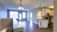 This well-appointed single-floor residence offers an open-concept layout that seamlessly connects the living, dining, and kitchen areas into one cohesive and functional space. Rich hardwood flooring flows throughout, lending warmth and continuity to the interior. The dining area is anchored by a classic multi-bulb chandelier, adding a timeless touch, while elegant pendant lights over the kitchen counter provide both task lighting and visual interest. The kitchen features white cabinetry, a dark countertop, and a tile backsplash, combining practicality with clean, contemporary style. Large windows and a set of glass-paneled doors flood the living and dining areas with generous natural light, creating a bright and airy atmosphere. The neutral gray wall tones complement the white trim and cabinetry, contributing to a cohesive and modern aesthetic. A doorway visible from the main living area suggests additional rooms beyond, offering further functional space. The overall ambiance is inviting and move-in ready, with a layout designed to support comfortable everyday living and easy entertaining.
