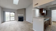 This single-floor, 2-bedroom, 2-bathroom residence offers a well-designed layout with notable architectural character throughout. The open-concept living and kitchen area serves as the heart of the home, highlighted by soaring vaulted ceilings, a skylight that fills the space with natural light, and a ceiling fan overhead. A black tile-surround fireplace with a white-painted mantel anchors the living room, while a sliding glass door opens to a balcony with outdoor views. The kitchen features rich, dark wood cabinetry, granite-style countertops with a breakfast bar peninsula, black appliances including a range and microwave, and durable wood-look flooring. A utility room with washer and dryer connections is accessible directly from the kitchen. The primary bedroom is generously proportioned and includes an en-suite bathroom equipped with a soaking tub and tile surround. The second bedroom is carpeted and bright, with its own adjacent full bathroom featuring a tub-shower combo, tile flooring, a wood vanity, and granite-style countertop. Both bedrooms include closets, and a hallway connects the private areas of the home. Neutral gray tones and plush carpeting throughout create a cohesive, move-in ready aesthetic.