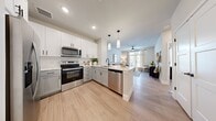 This well-designed one-bedroom, one-bathroom unit features a thoughtfully organized open-concept layout with light wood-look flooring and a neutral color palette throughout. The kitchen is a standout feature, equipped with stainless steel appliances including a French door refrigerator, electric range, microwave, and dishwasher. White and gray shaker-style cabinetry extends to the ceiling, complemented by a white subway tile backsplash and light quartz countertops. A peninsula island with seating provides additional prep space and casual dining options, while pendant lighting adds a modern touch overhead. The kitchen flows seamlessly into the living area, which benefits from natural light through large windows and a ceiling fan. The generously sized primary bedroom accommodates a king-size bed comfortably and features multiple windows with blinds and sheer curtains. An adjoining walk-in closet offers built-in shelving and hanging rods for organized storage. The full bathroom features gray shaker vanity cabinetry, a quartz countertop, a soaking tub with shower, and matte black hardware accents, creating a cohesive, contemporary aesthetic throughout the space.