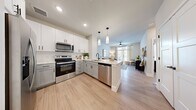 This well-designed one-bedroom, one-bathroom unit features a thoughtfully organized open-concept layout with light wood-look flooring and a neutral color palette throughout. The kitchen is a standout feature, equipped with stainless steel appliances including a French door refrigerator, electric range, microwave, and dishwasher. White and gray shaker-style cabinetry extends to the ceiling, complemented by a white subway tile backsplash and light quartz countertops. A peninsula island with seating provides additional prep space and casual dining options, while pendant lighting adds a modern touch overhead. The kitchen flows seamlessly into the living area, which benefits from natural light through large windows and a ceiling fan. The generously sized primary bedroom accommodates a king-size bed comfortably and features multiple windows with blinds and sheer curtains. An adjoining walk-in closet offers built-in shelving and hanging rods for organized storage. The full bathroom features gray shaker vanity cabinetry, a quartz countertop, a soaking tub with shower, and matte black hardware accents, creating a cohesive, contemporary aesthetic throughout the space.