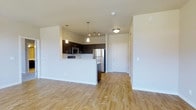 This single-floor space showcases a clean, modern design with an open-concept layout that seamlessly connects the kitchen and living areas. The kitchen is a standout feature, outfitted with rich, dark espresso cabinetry that contrasts beautifully against crisp white walls and light hardwood-style flooring that extends throughout the unit. Stainless steel appliances — including a full-size refrigerator and microwave — lend a contemporary, polished feel, while a peninsula-style breakfast bar provides a practical and social dining surface. Pendant lighting hangs above the kitchen, adding warm, focused illumination that complements the flush ceiling fixtures in the adjacent spaces. The open living area is bright and airy, benefiting from the clean lines and neutral color palette that create a welcoming, versatile atmosphere. A clearly defined entryway with a solid white door offers a structured transition into the main living space. The layout is efficient yet comfortable, with thoughtful design details that balance functionality and style. Light-toned flooring and white walls ensure the space feels open and well-lit, making it easy to personalize and furnish to a variety of tastes.