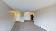 This single-floor unit offers an open-concept layout that connects the living area to the kitchen, creating a functional and cohesive living space. The living area is finished with wall-to-wall carpet in a neutral tone, complemented by light grey walls and white baseboards that lend a clean, understated aesthetic throughout. A textured popcorn ceiling adds dimension overhead, while a ceiling fan positioned between the living and kitchen areas provides both air circulation and ambient lighting.

The entryway is clearly defined near one end of the space, featuring a white paneled door with overhead lighting, providing a welcoming transition into the unit. A baseboard heater along the exterior wall offers an efficient heating solution for the living space.

The kitchen is partially visible from the main living area and features black appliances, including a full-sized refrigerator, set against light-colored cabinetry. The kitchen flooring transitions from the carpeted living area, helping to visually distinguish the two spaces. Overall, the unit presents a straightforward, well-maintained interior with a practical and open design suited for comfortable everyday living.
