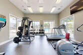 This fitness center is a compact yet comprehensively outfitted space designed to support a full range of workout routines. The room is flooded with natural light through large sliding and picture windows positioned along multiple walls, offering pleasant views of an outdoor pool area and surrounding greenery — creating an energizing atmosphere for exercise.

The flooring consists of durable, neutral-toned gym tiles suited for heavy equipment use. A full-wall mirror spans one side of the room, visually expanding the space and allowing users to monitor their form during workouts. Flush-panel fluorescent lighting is evenly distributed across the white ceiling, ensuring bright, consistent illumination throughout.

Cardio equipment includes treadmills, an elliptical trainer, and a Precor elliptical/cycling machine. For strength training, the space features a Magnum multi-function cable machine with weight plates, a three-tier dumbbell rack stocked with a wide range of free weights, and a red adjustable workout bench. A wall-mounted flat-screen television is positioned above the cardio zone for entertainment during workouts. A hand sanitizer dispenser is also mounted on the wall, reflecting attention to cleanliness. The overall ambiance is clean, functional, and purpose-built for an effective fitness experience.