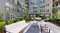 This single-floor space is defined by two distinct environments that together shape its overall character. The interior bathroom is compact and straightforward, featuring a wood-cabinet vanity with an integrated sink, chrome fixtures, and a framed mirror with warm wood detailing. A metal over-toilet shelving unit provides additional vertical storage, and neutral wall tones with gold-toned hardware accents lend a classic, understated feel to the space.

The most striking feature associated with this property is its access to a beautifully designed courtyard. Enclosed by a multi-story building with gray horizontal siding, private balconies, and large windows, the outdoor area is thoughtfully landscaped with raised garden beds, ornamental shrubs, young trees, and lush greenery. The hardscape patio is generously sized, furnished with contemporary outdoor seating arrangements including white sofas, armchairs, and low coffee tables. A shaded seating area with an umbrella is also visible, offering a cooler retreat. Natural light fills the courtyard throughout the day, and the surrounding architecture creates an intimate, sheltered atmosphere that balances open-air freshness with a sense of privacy.