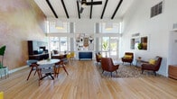 This single-floor space showcases a striking open-concept layout defined by soaring vaulted ceilings with exposed dark wood beams and abundant natural light streaming in through floor-to-ceiling clerestory windows. The wood-look tile flooring runs throughout, lending warmth and continuity across the entire space.

The main gathering area serves as the centerpiece, featuring a curated seating arrangement of rich brown leather armchairs accented with mustard yellow throw pillows, arranged over a patterned area rug. A statement gallery wall displays an eclectic mix of framed art, woven wall hangings, and decorative objects above a sleek navy credenza, adding a creative, personalized touch.

Adjacent to the lounge area is a functional workstation zone, anchored by dark wood desks with computer monitors and vibrant yellow accent chairs. A textured, warm-toned decorative wall adds depth and character behind the work area. Nearby, a casual dining arrangement with a round white table and mid-century modern chairs provides a collaborative setting.

The entry area features large glass double doors that invite natural light and outdoor views, flanked by a reception-style counter with built-in cabinetry. A separate seating nook, partially divided by a decorative lattice screen, offers a more private retreat. Together, these zones create a welcoming, functional, and stylish environment suited for both social interaction and focused work.