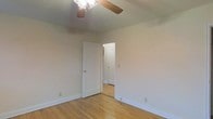 This single-floor space presents a straightforward and functional design. The room visible in the image features light hardwood flooring that adds warmth to the neutral, beige-toned walls. A ceiling fan with wooden blades and an integrated light fixture is mounted overhead, providing both air circulation and ambient lighting. White baseboards frame the perimeter of the room, offering a clean contrast against the flooring. An open doorway connects to an adjacent room or hallway, suggesting a practical, flowing layout between spaces. The arched transition visible beyond the doorway hints at subtle architectural detailing throughout. The overall ambiance is understated and move-in ready, with a simple palette that allows for versatile interior styling.